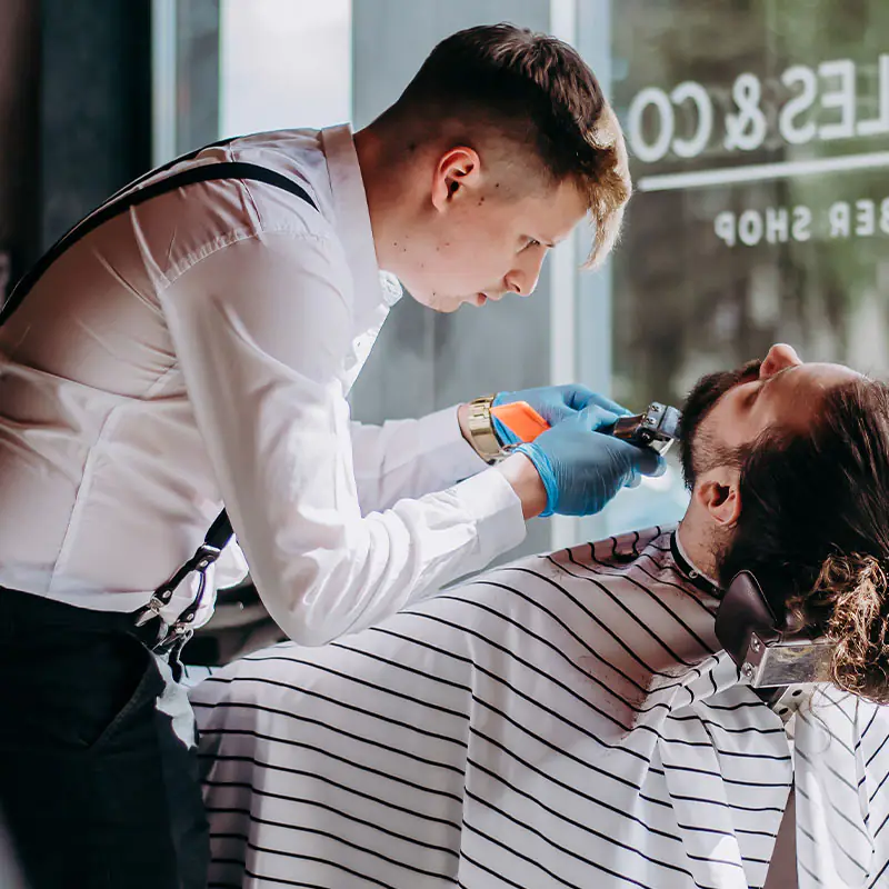 Barber Charles & Co podczas strzyżenia brody — koszula z szelkami, niebieska rękawica, działanie precyzyjne